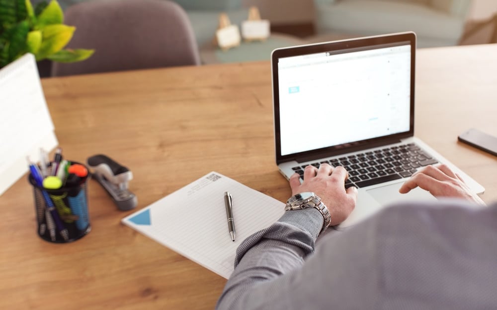 Man working on his laptop in office, blurred notepad, markers and calendar at desk, view from behind, detail on hand and wristwatch